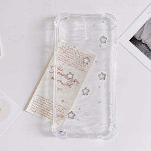 May include: Clear phone case with a decorative design. The case features embedded star-shaped embellishments and iridescent glitter. A handwritten note with the words "I love you so much" is partially visible behind the case.