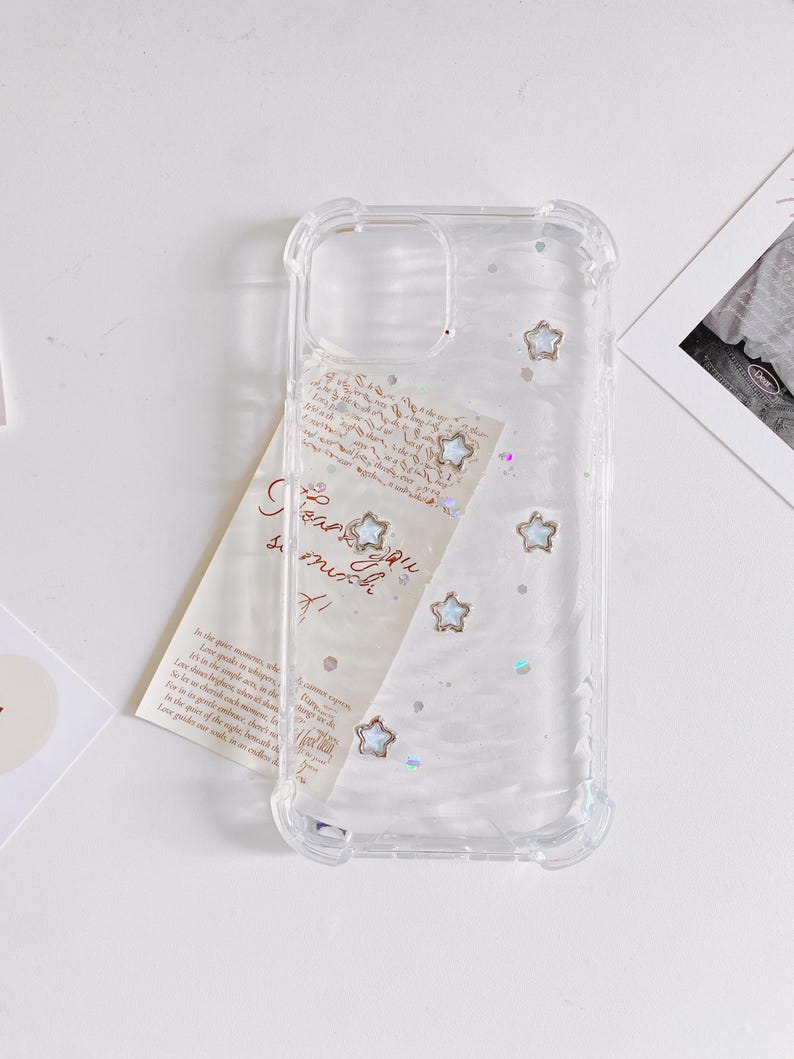 May include: Clear phone case with a decorative design. The case features embedded star-shaped embellishments and iridescent glitter. A handwritten note with the words "I love you so much" is partially visible behind the case.