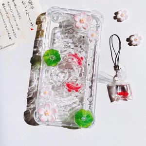 Pu&ograve; includere: Custodia trasparente per telefono decorata con pesci koi rossi, fiori rosa e foglie di ninfea verdi. La custodia ha un effetto simile all'acqua con glitter. &Egrave; presente anche un piccolo ciondolo a forma di pesce e ulteriori abbellimenti floreali.