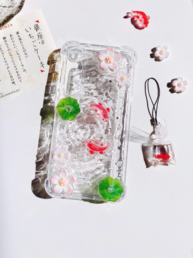 Pu&ograve; includere: Custodia trasparente per telefono decorata con pesci koi rossi, fiori rosa e foglie di ninfea verdi. La custodia ha un effetto simile all'acqua con glitter. &Egrave; presente anche un piccolo ciondolo a forma di pesce e ulteriori abbellimenti floreali.