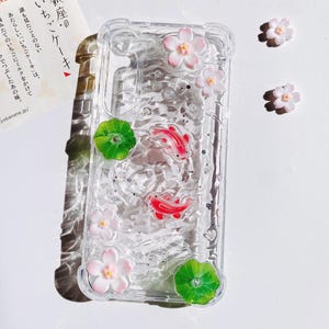 Pu&ograve; includere: Custodia trasparente per telefono decorata con pesci koi rossi, fiori rosa e bianchi e foglie di ninfea verdi. La custodia ha un aspetto strutturato simile all'acqua con glitter incorporati. Ulteriori elementi decorativi sono sparsi.