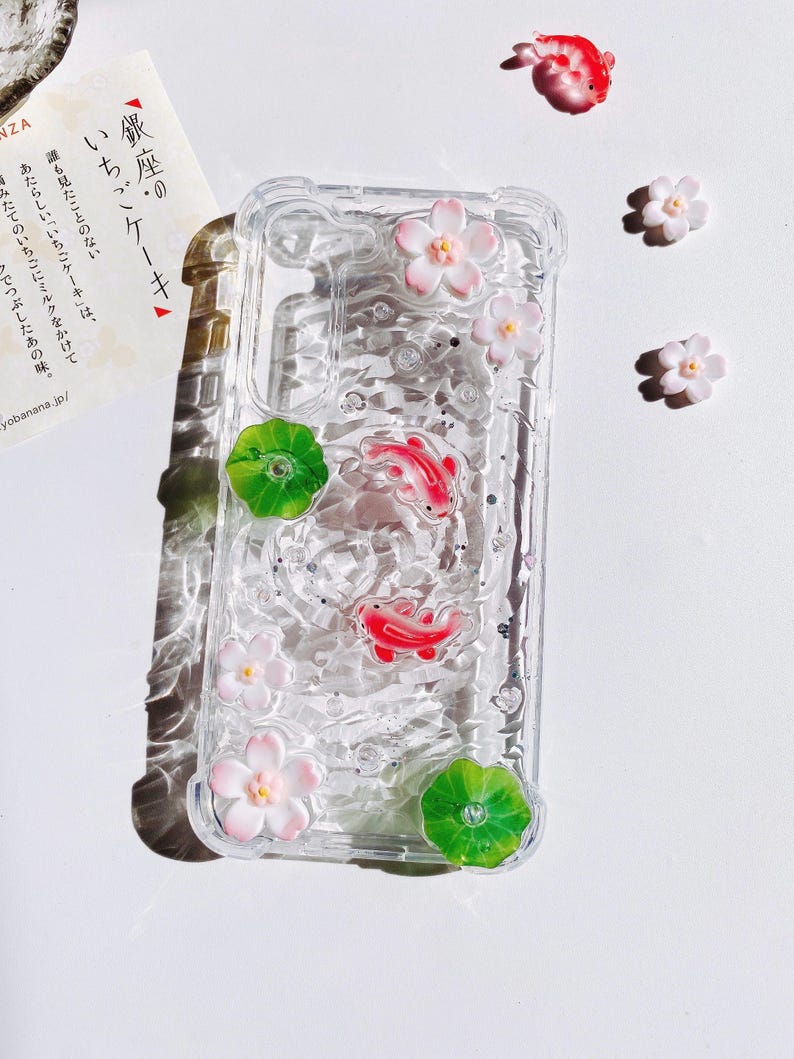Pu&ograve; includere: Custodia trasparente per telefono decorata con pesci koi rossi, fiori rosa e bianchi e foglie di ninfea verdi. La custodia ha un aspetto strutturato simile all'acqua con glitter incorporati. Ulteriori elementi decorativi sono sparsi.
