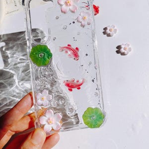 Pu&ograve; includere: Custodia trasparente per telefono con pesci koi rosa, fiori bianchi e rosa, foglie di ninfea verdi e glitter argentati. La custodia &egrave; tenuta in mano. La custodia ha il testo giapponese "座の・ちごケ".
