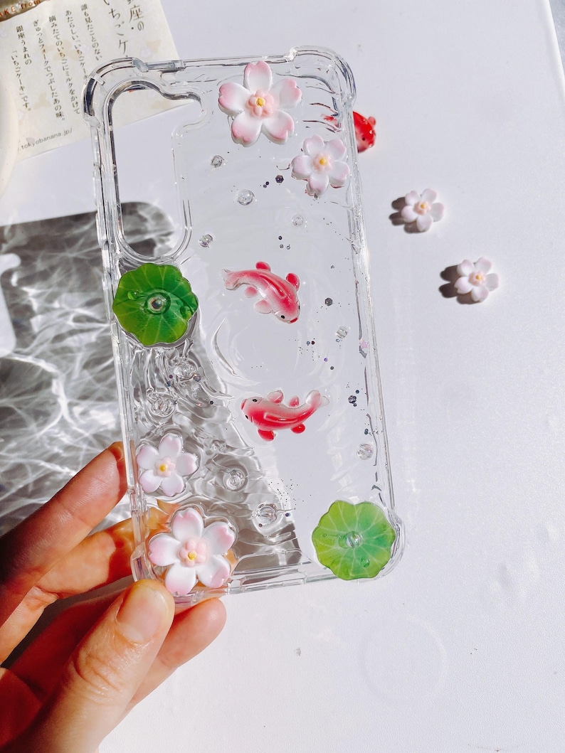 Pu&ograve; includere: Custodia trasparente per telefono con pesci koi rosa, fiori bianchi e rosa, foglie di ninfea verdi e glitter argentati. La custodia &egrave; tenuta in mano. La custodia ha il testo giapponese "座の・ちごケ".