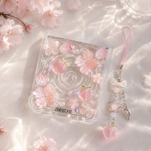 May include: Clear phone case decorated with pink cherry blossom and peach designs. A matching pink charm with a bow, flowers, and a peach pendant hangs from the case. The text "JAMAECAS" is visible on the case.