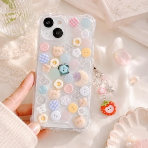 May include: Clear phone case decorated with colorful buttons, flowers, and bear shapes. The case has a charm with beads and a strawberry. The case is designed for a smartphone.