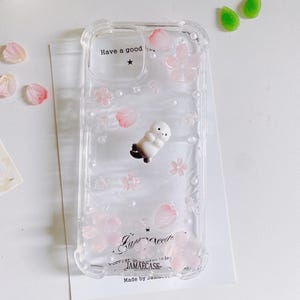 Może przedstawiać: Przezroczyste etui na telefon z dekoracyjnym wzorem. Etui posiada małą białą figurkę foki, r&oacute;żowe kwiatowe akcenty oraz tekst "Have a good" i "Fair reeds". Etui jest wykonane przez JAMAECASE.