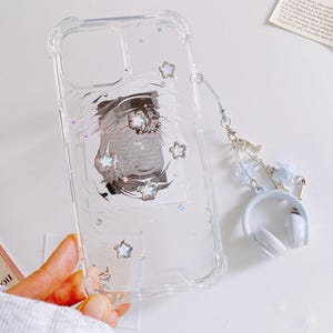 May include: Clear phone case with a protective bumper, featuring embedded silver star accents and a swirling design. Includes a matching charm with a miniature white headphone pendant and a light blue strap.