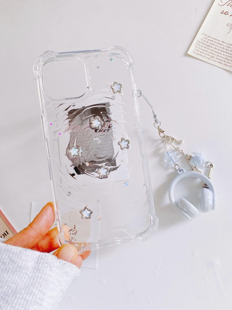 May include: Clear phone case with a protective bumper, featuring embedded silver star accents and a swirling design. Includes a matching charm with a miniature white headphone pendant and a light blue strap.