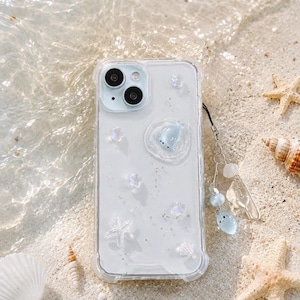 May include: Clear phone case with a sea-themed design. The case features a clear background with embedded white starfish and iridescent glitter. A small blue seal charm and a matching wrist strap with seashell and bead accents complete the design. The case is resting on a sandy beach.