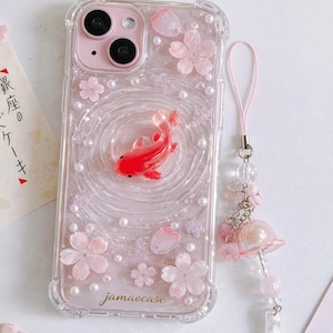 以下が含まれることがあります： ピンク色のカメラ部分が付いた透明な携帯電話ケース。赤い鯉、ピンクの花、真珠が特徴です。ケースには、チャームが付いたお揃いのピンク色のストラップが付いています。「jamaecase」のブランド名が表示されています。