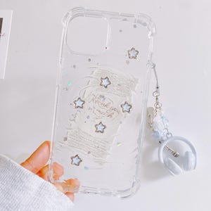 May include: Clear phone case with star and glitter accents. The case has a white design with the words "Thank you much" and several star-shaped embellishments. A white headphone charm is attached.