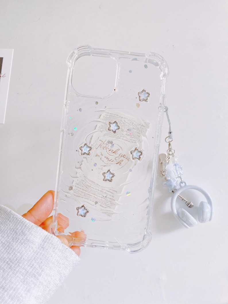 May include: Clear phone case with star and glitter accents. The case has a white design with the words "Thank you much" and several star-shaped embellishments. A white headphone charm is attached.