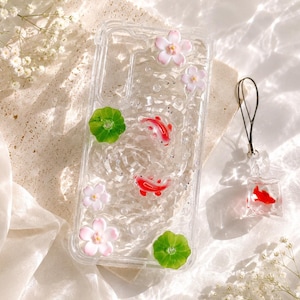 Pu&ograve; includere: Custodia trasparente per telefono con pesci koi rossi, fiori rosa e foglie di ninfea verdi. Un ciondolo a forma di pesce koi abbinato &egrave; appeso a un cordino nero. La custodia ha un aspetto glitterato e acquatico.