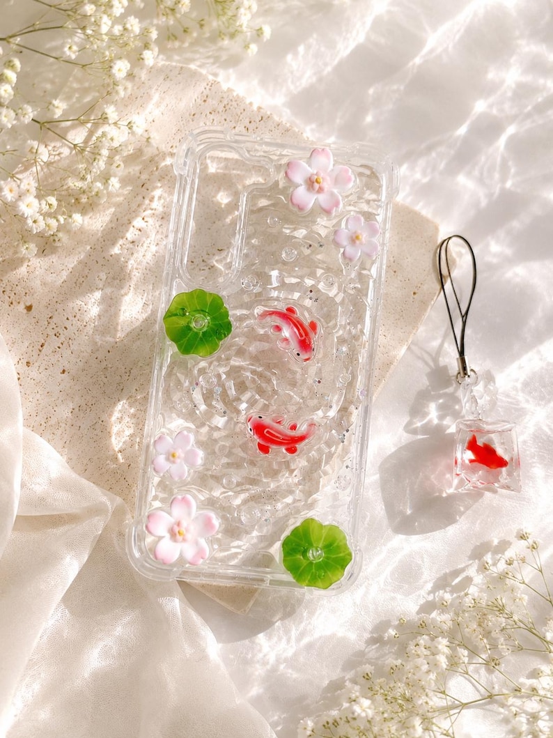 Pu&ograve; includere: Custodia trasparente per telefono con pesci koi rossi, fiori rosa e foglie di ninfea verdi. Un ciondolo a forma di pesce koi abbinato &egrave; appeso a un cordino nero. La custodia ha un aspetto glitterato e acquatico.
