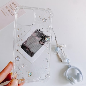 May include: Clear phone case with glitter, stars, and a small planet charm. The case has a decorative charm with a microphone, stars, and a headset. A small card with text is visible behind the case.