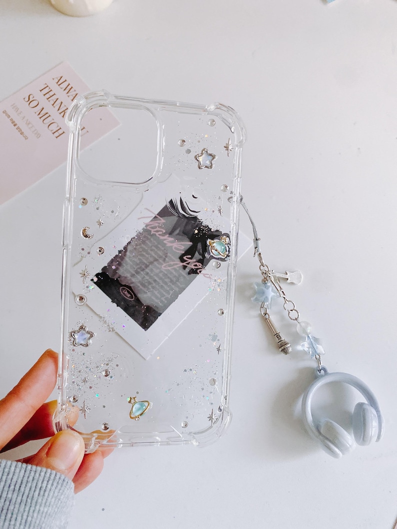 May include: Clear phone case with glitter, stars, and a small planet charm. The case has a decorative charm with a microphone, stars, and a headset. A small card with text is visible behind the case.