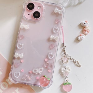 May include: A clear phone case with a pink background and white bows, hearts, and flowers. The case has a peach charm attached to it.