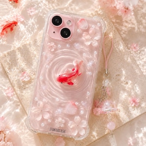 以下が含まれることがあります： ピンク色の内装の透明な携帯電話ケース。3Dの金魚と花のアクセントが特徴です。ケースにはピンク色のストラップとチャームが付いています。「JAMAECASE」の文字がケースにプリントされています。