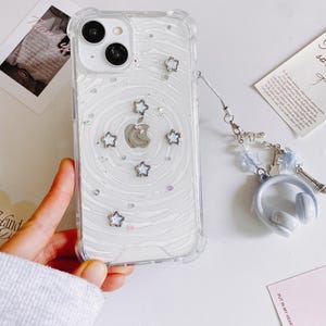 May include: Clear phone case with a white swirl design, silver star accents, and a silver Apple logo. The case has a clear bumper and a charm with headphones and a small Eiffel Tower. The phone case is held by a person.