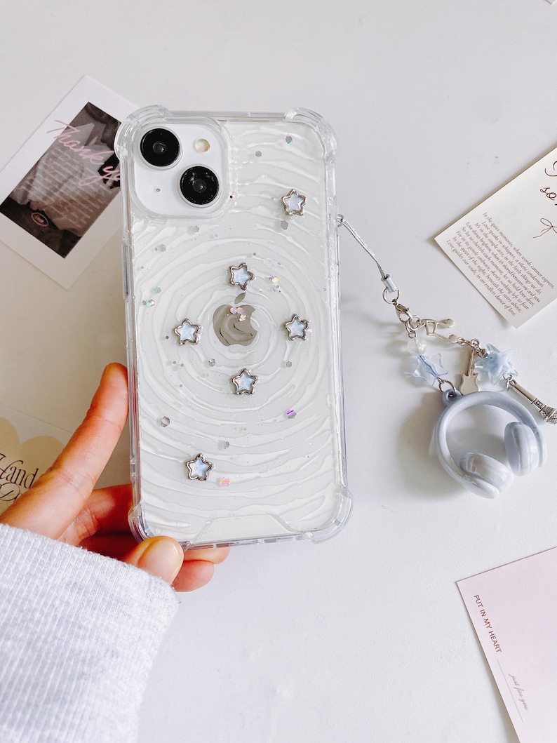 May include: Clear phone case with a white swirl design, silver star accents, and a silver Apple logo. The case has a clear bumper and a charm with headphones and a small Eiffel Tower. The phone case is held by a person.
