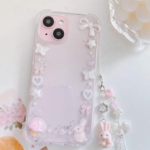 May include: A clear phone case with a pink interior, decorated with white bows, stars, hearts, and butterflies. The case features a small pink mushroom and a bunny. A charm with a bunny, flowers, and bows hangs from the case.