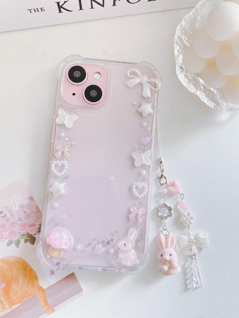 May include: A clear phone case with a pink interior, decorated with white bows, stars, hearts, and butterflies. The case features a small pink mushroom and a bunny. A charm with a bunny, flowers, and bows hangs from the case.