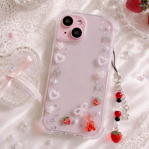 May include: A pink phone case with a clear border, decorated with white pearls, hearts, bows, stars, and red strawberry and gummy bear charms. A black lanyard with red, black, and white beads and a strawberry charm is attached.