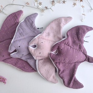 May include: Four plush baby toys in the shape of stingrays, in shades of purple and pink. Each toy has a white lining and is dotted with gold-colored sparkles. The toys are arranged on a white surface.