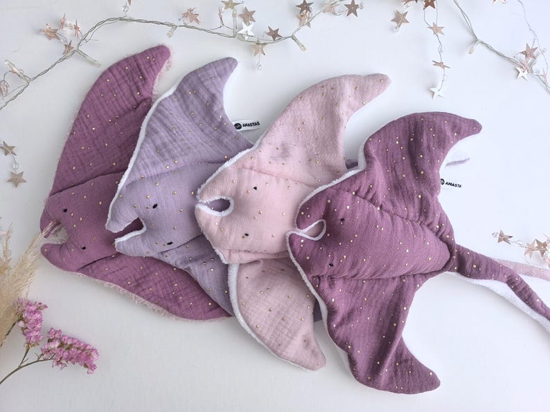 May include: Four plush baby toys in the shape of stingrays, in shades of purple and pink. Each toy has a white lining and is dotted with gold-colored sparkles. The toys are arranged on a white surface.
