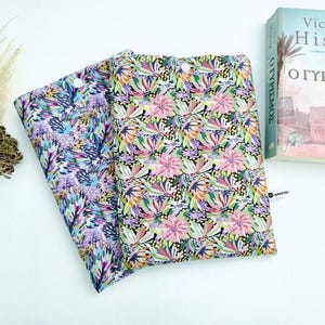 Op de afbeelding: Twee stoffen boekomslagen met bloempatronen. Eén omslag heeft een paars en blauw botanisch ontwerp, de andere een mix van roze, groene en gele bloemen op een zwarte achtergrond. Een boek is zichtbaar op de achtergrond.