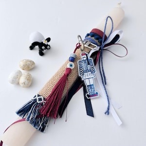 May include: A decorative candle with a burlap wrap, adorned with a tassel, ribbons, and a keychain featuring a traditional Greek soldier. Additional decorations include a small black and white sheep figurine and speckled eggs.