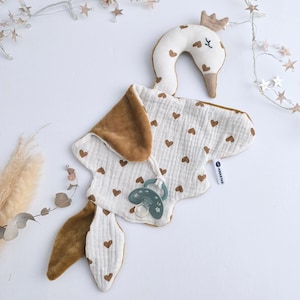 May include: A baby gift set featuring a swan-shaped plush toy with a crown, a bib with brown heart patterns, and a pacifier. The bib and swan are white with brown accents. The pacifier is teal.