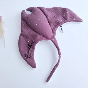 May include: A purple manta ray-shaped baby toy with gold speckles. The toy has a long tail and the name "Coraline" embroidered in black. The toy is made of soft fabric and has a white underside.