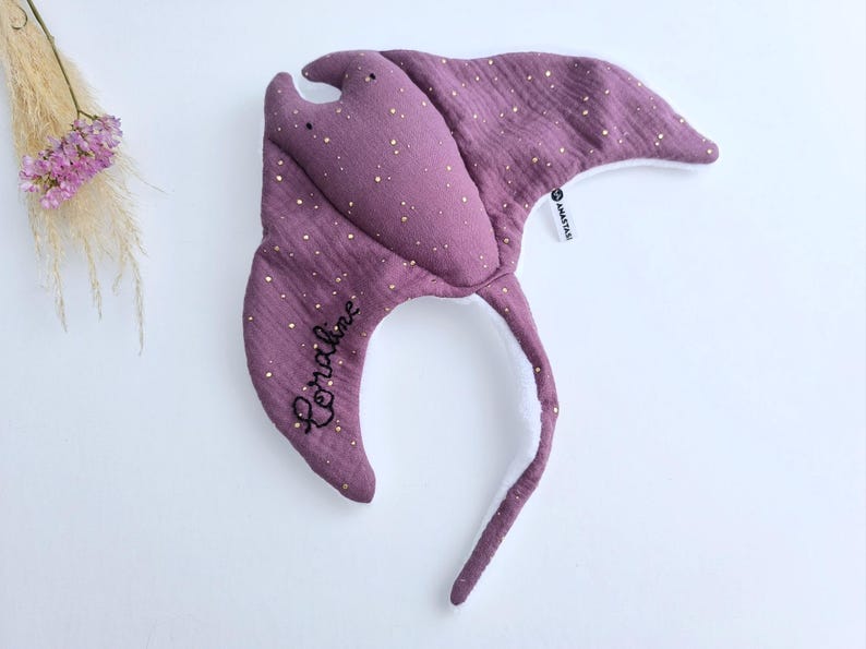 May include: A purple manta ray-shaped baby toy with gold speckles. The toy has a long tail and the name "Coraline" embroidered in black. The toy is made of soft fabric and has a white underside.