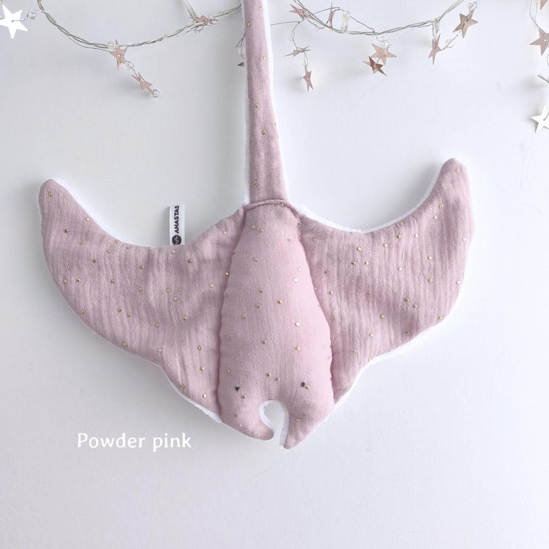 May include: A powder pink, plush stingray-shaped baby mobile with gold-colored accents. The mobile is made of soft fabric and has a white underside. The text "ANASTAS" is visible on the left wing. The text "Powder pink" is at the bottom.
