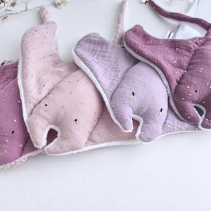 May include: A collection of baby bibs in various shades of purple and pink, shaped like manta rays. Each bib is adorned with small, gold-colored embellishments and has a white trim. The bibs are made of soft, textured fabric.