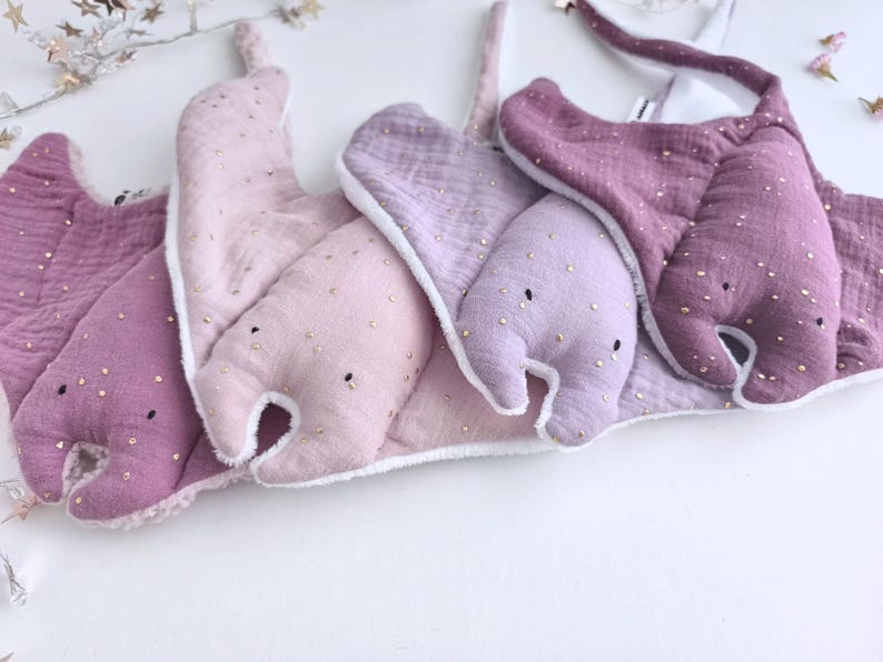 May include: A collection of baby bibs in various shades of purple and pink, shaped like manta rays. Each bib is adorned with small, gold-colored embellishments and has a white trim. The bibs are made of soft, textured fabric.