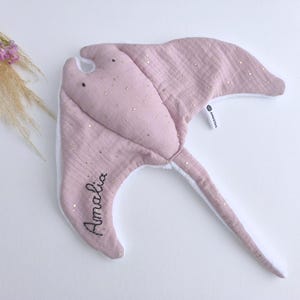 May include: A soft, pink manta ray-shaped baby toy with gold glitter accents. The toy has a white underside and a long tail. The name "Amalia" is embroidered on one wing. Decorative flowers are in the background.