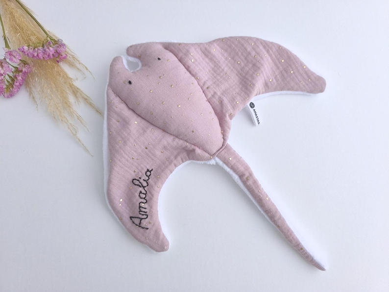 May include: A soft, pink manta ray-shaped baby toy with gold glitter accents. The toy has a white underside and a long tail. The name "Amalia" is embroidered on one wing. Decorative flowers are in the background.