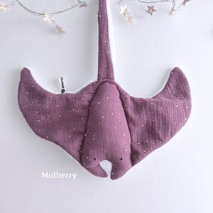 May include: A soft, purple stuffed manta ray with gold speckles. The toy has a long tail and wide wings, with a white underside. The word "Mulberry" is printed at the bottom. A string of star-shaped lights hangs above the toy.