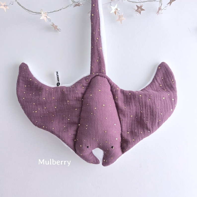 May include: A soft, purple stuffed manta ray with gold speckles. The toy has a long tail and wide wings, with a white underside. The word "Mulberry" is printed at the bottom. A string of star-shaped lights hangs above the toy.