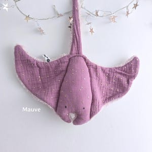 May include: Mauve-colored plush stingray toy with gold-colored accents. The toy has a soft, textured appearance and a whimsical design. The word "Mauve" is printed on the bottom left of the image. The toy is suspended from a string of star-shaped lights.