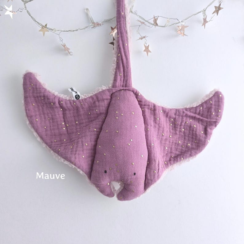 May include: Mauve-colored plush stingray toy with gold-colored accents. The toy has a soft, textured appearance and a whimsical design. The word "Mauve" is printed on the bottom left of the image. The toy is suspended from a string of star-shaped lights.
