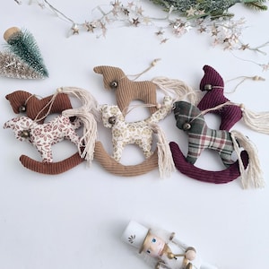 Puede incluir: Una colección de cinco adornos de tela con forma de caballo balancín que presentan diversas texturas y estampados, como pana lisa, motivos florales y cuadros. Cada caballo está decorado con un pequeño cascabel, una cola de borla y un cordel.