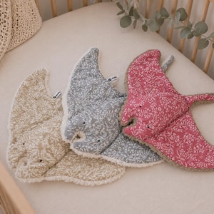Op de afbeelding: Drie zachte speelgoedjes in de vorm van een manta rog in een wieg. Eén is beige, één is grijs en één is roze. Elk heeft een bloemenpatroon en een zachte, getextureerde rand. Het speelgoed ligt op een wit laken.