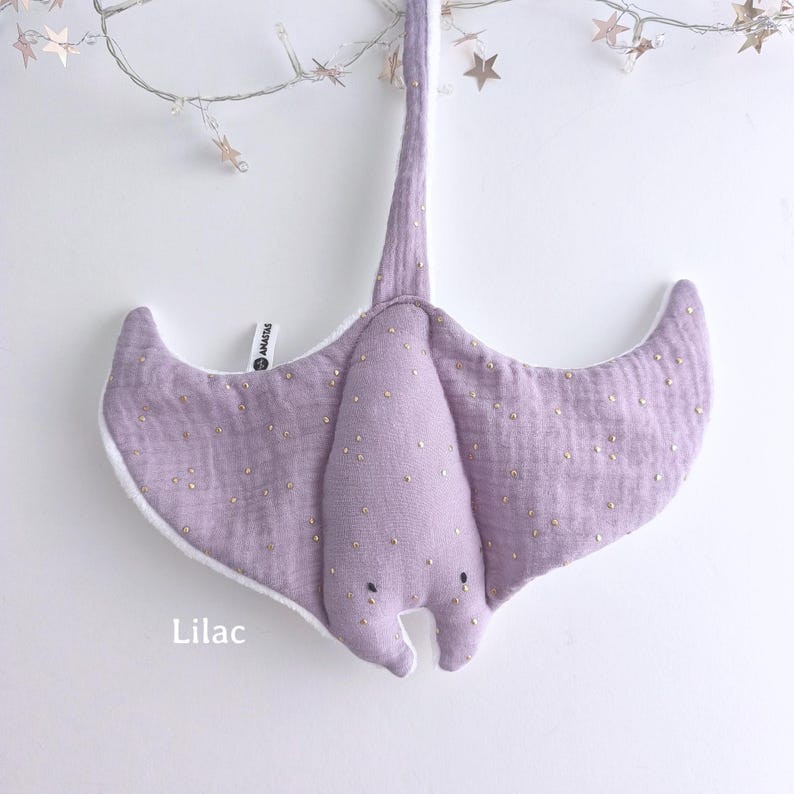May include: A lilac-colored stuffed stingray toy with gold-colored speckles. The toy has a white border and a long, slender tail. The word "Lilac" is printed at the bottom of the image. A string of lights with star-shaped ornaments hangs above the toy.