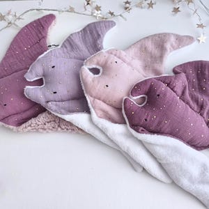 May include: Four plush baby toys shaped like manta rays in shades of purple, pink, and white. Each toy has a soft, textured fabric with gold accents. The toys are arranged on a white surface, with a string of star-shaped lights in the background.