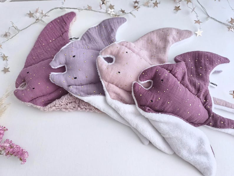 May include: Four plush baby toys shaped like manta rays in shades of purple, pink, and white. Each toy has a soft, textured fabric with gold accents. The toys are arranged on a white surface, with a string of star-shaped lights in the background.