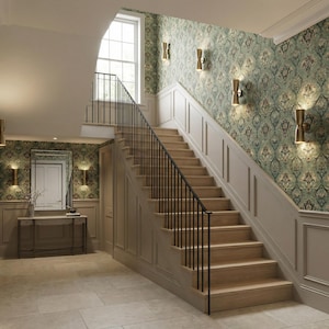 May include: An interior shot of a grand staircase with light wood steps and a black iron railing. The walls are painted in a neutral color with decorative paneling and wallpaper with a floral pattern. Gold sconces provide lighting.
